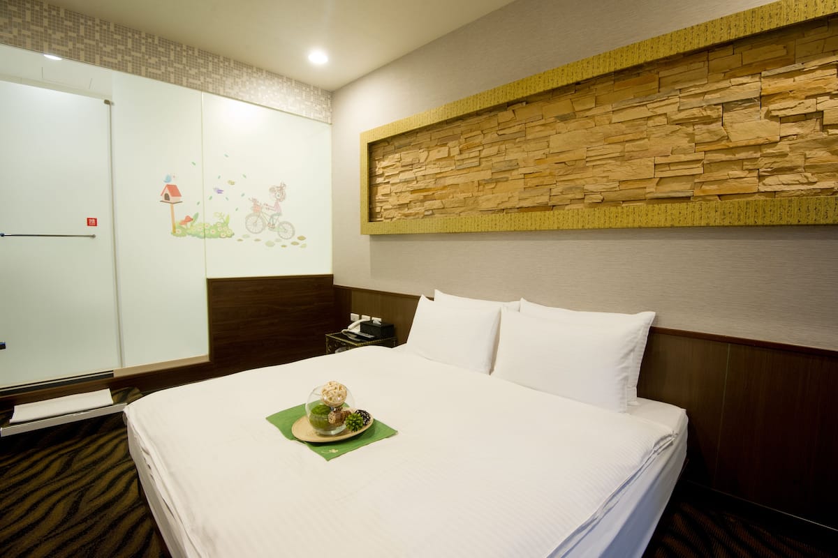 A hotel room with a neatly made bed, a wall-mounted artwork, a wooden headboard, and a small table with a decorative item.