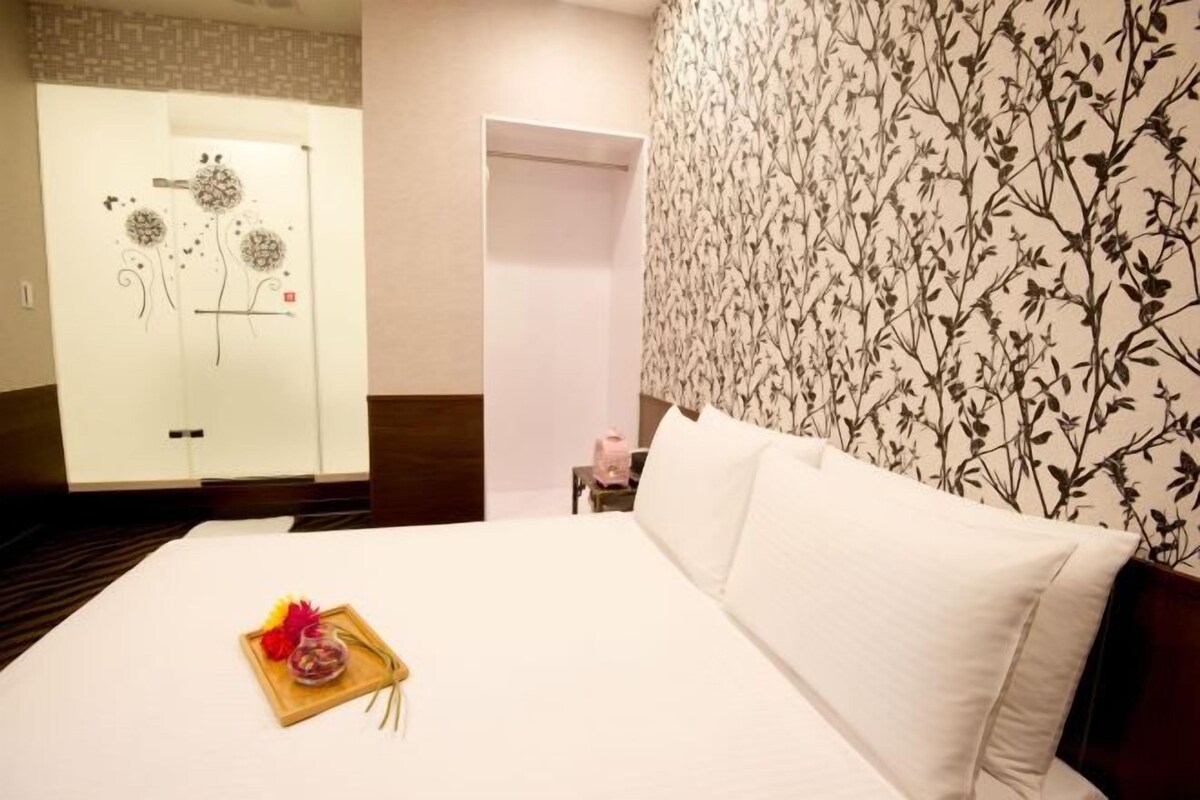 A hotel room with a bed, a nightstand, a bathroom with a shower, and a wall with a floral pattern.