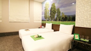 A hotel room with two beds, a green floral arrangement on the left bed, a nightstand with a picture frame and glasses, and a mural of trees on the wall.