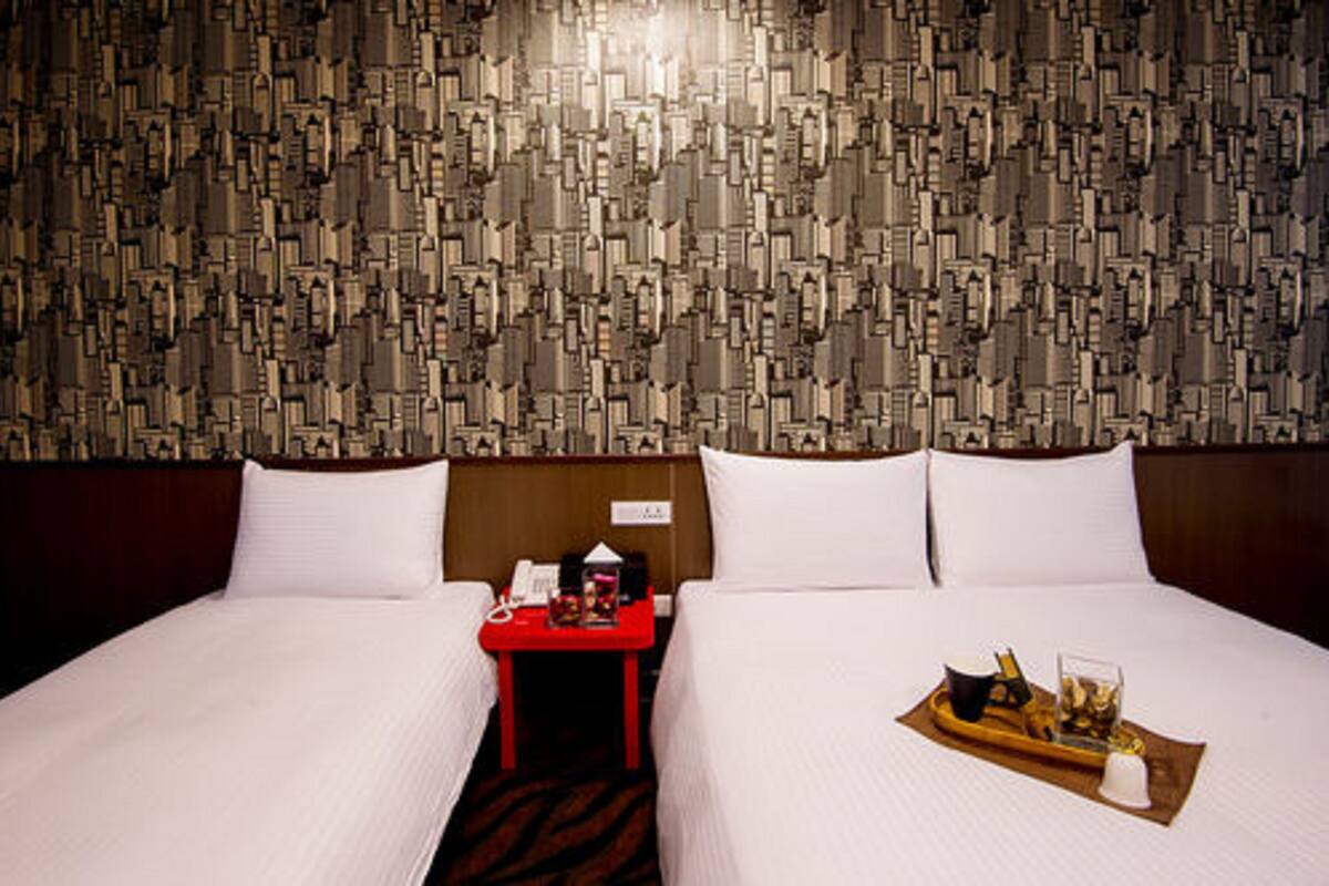 A hotel room with a patterned wall, two beds, a red nightstand, and a tray with items on the bed.
