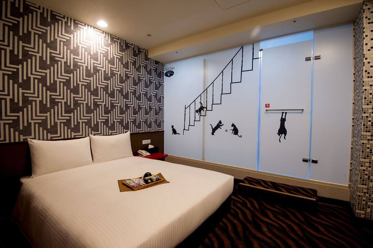 A hotel room with a bed, a nightstand, a tray with items, a patterned wall, a staircase with cat silhouettes, and a tiled wall.