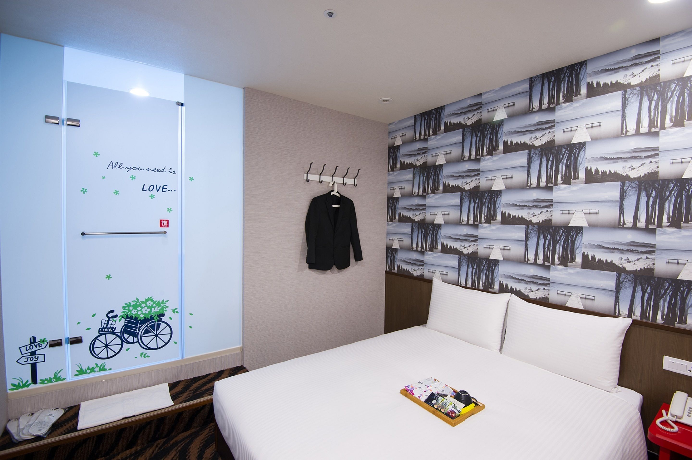 A hotel room with a bed, a coat rack with a black jacket, a wall with a forest wallpaper, and a door with a bicycle and 'LOVE' decoration.