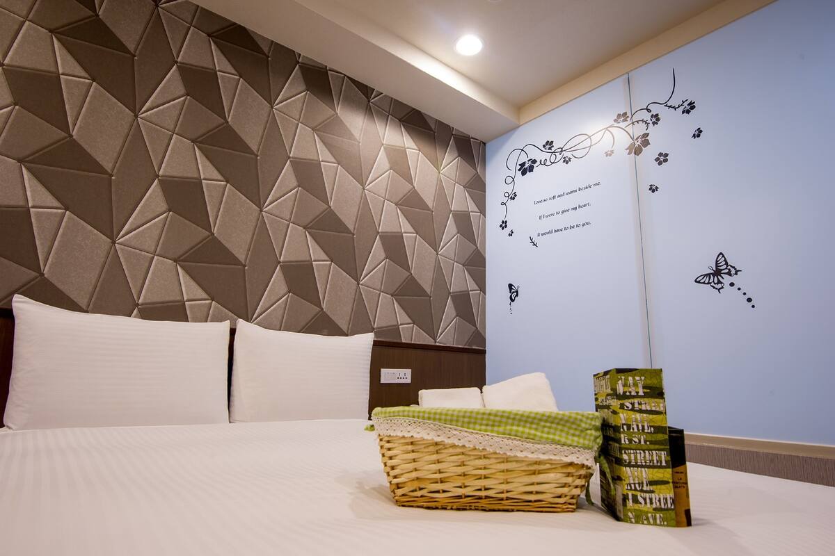 A hotel room with a geometric-patterned headboard, a neatly made bed with white linens, a decorative wall with floral and butterfly decals, and a stack of books on the side.