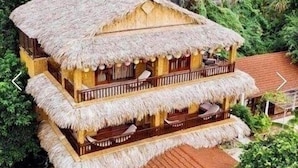 A multi-story building with a thatched roof and wooden balconies, surrounded by lush greenery and rocky terrain. - Mua Caves Ecolodge (Hoa Lu)