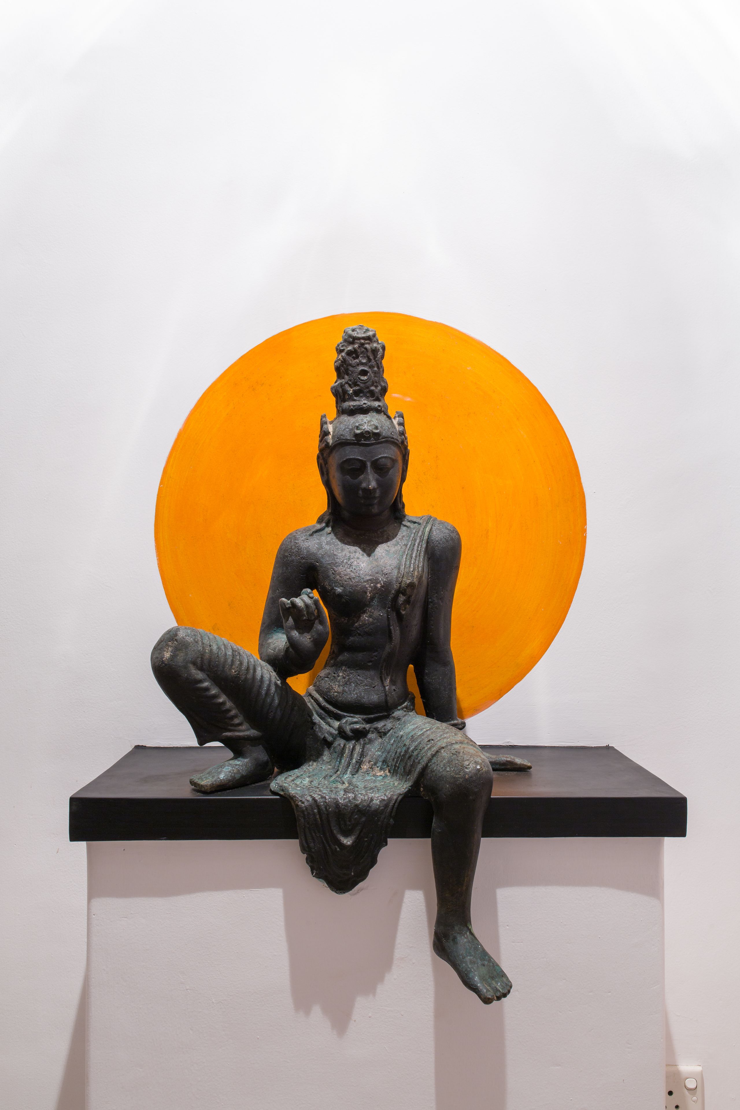 Een beeld van een zittende figuur op een zwart voetstuk, voor een witte muur met een grote oranje cirkel erachter.