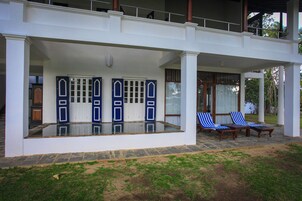 A two-story building with a covered patio area featuring a glass-enclosed pool, two lounge chairs, and a set of blue and white doors.