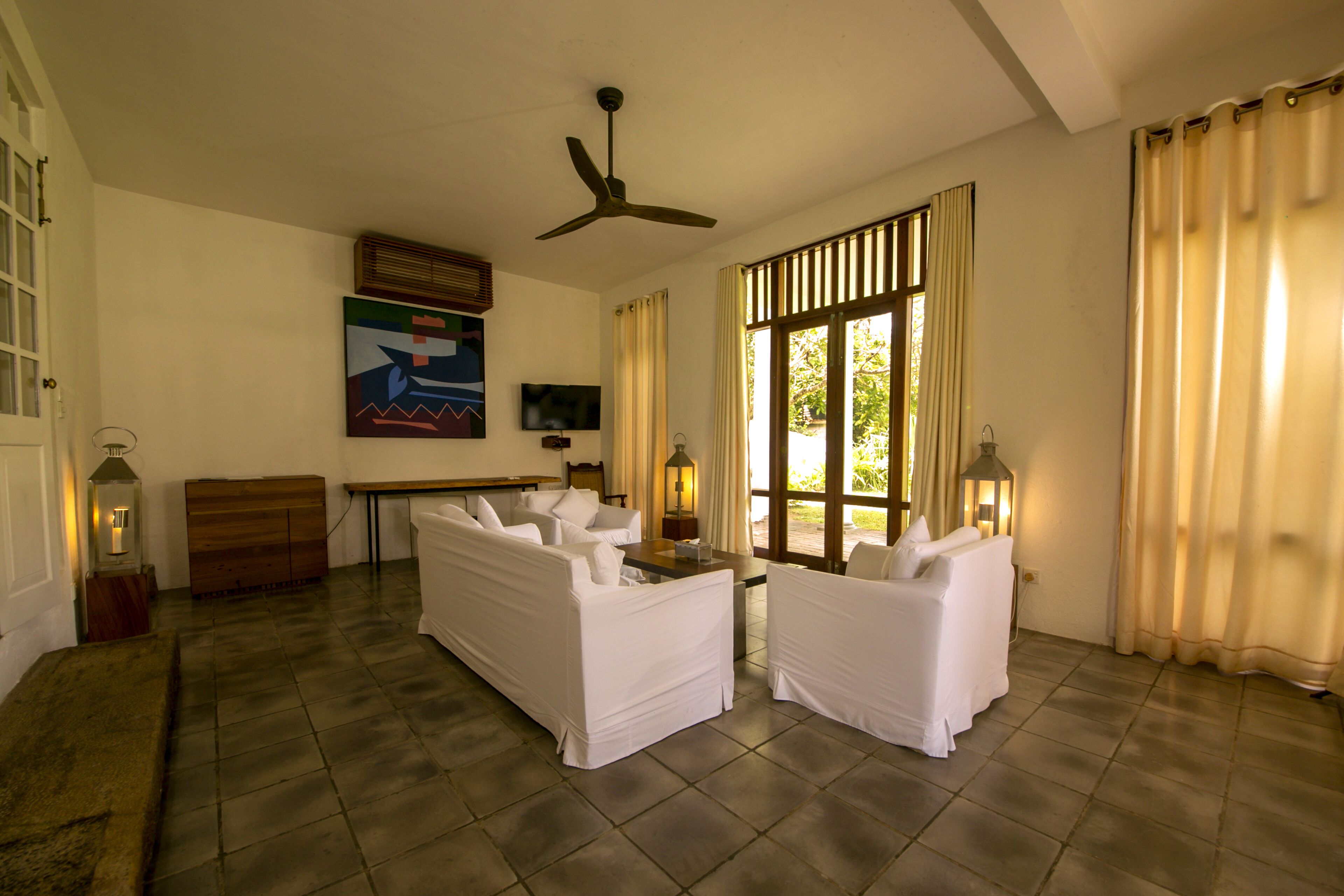 A spacious living room with a ceiling fan, white sofas, a wooden cabinet, and a TV on the wall.