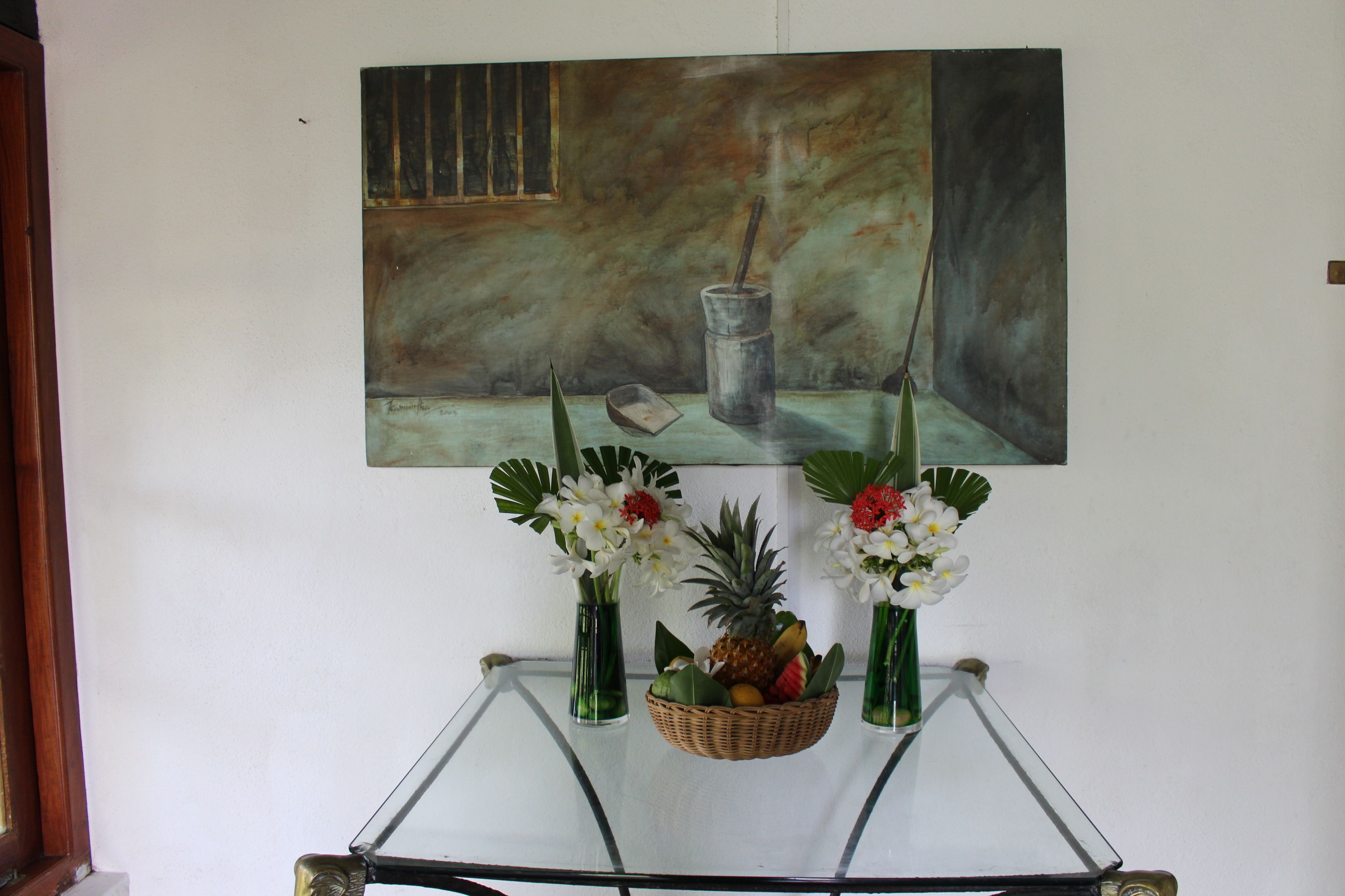 Una habitación con una mesa de vidrio que muestra un cuadro, dos jarrones con flores y una cesta de mimbre con frutas.