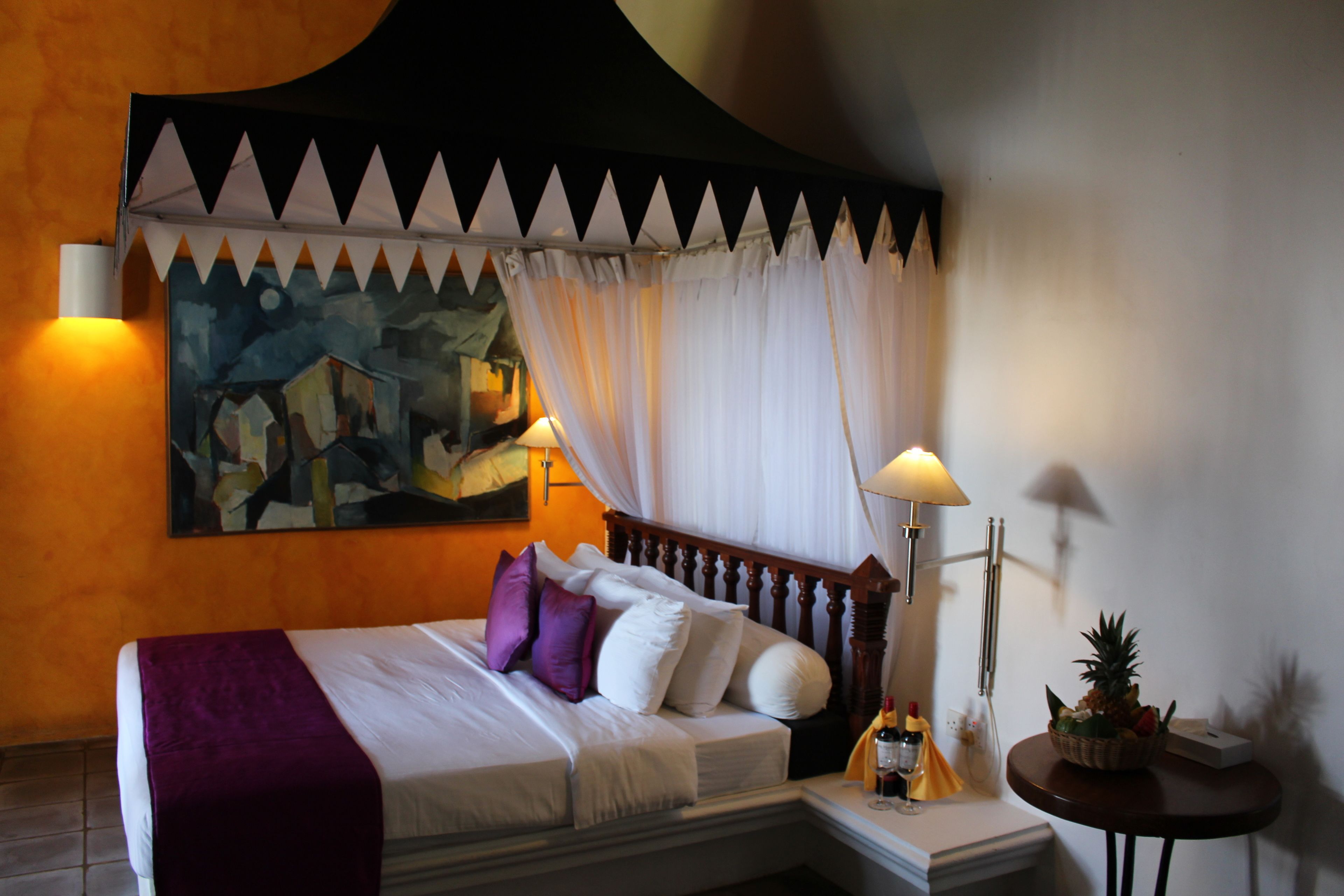 A hotel room with a bed, a nightstand, a lamp, a fruit bowl, and a painting on the wall.