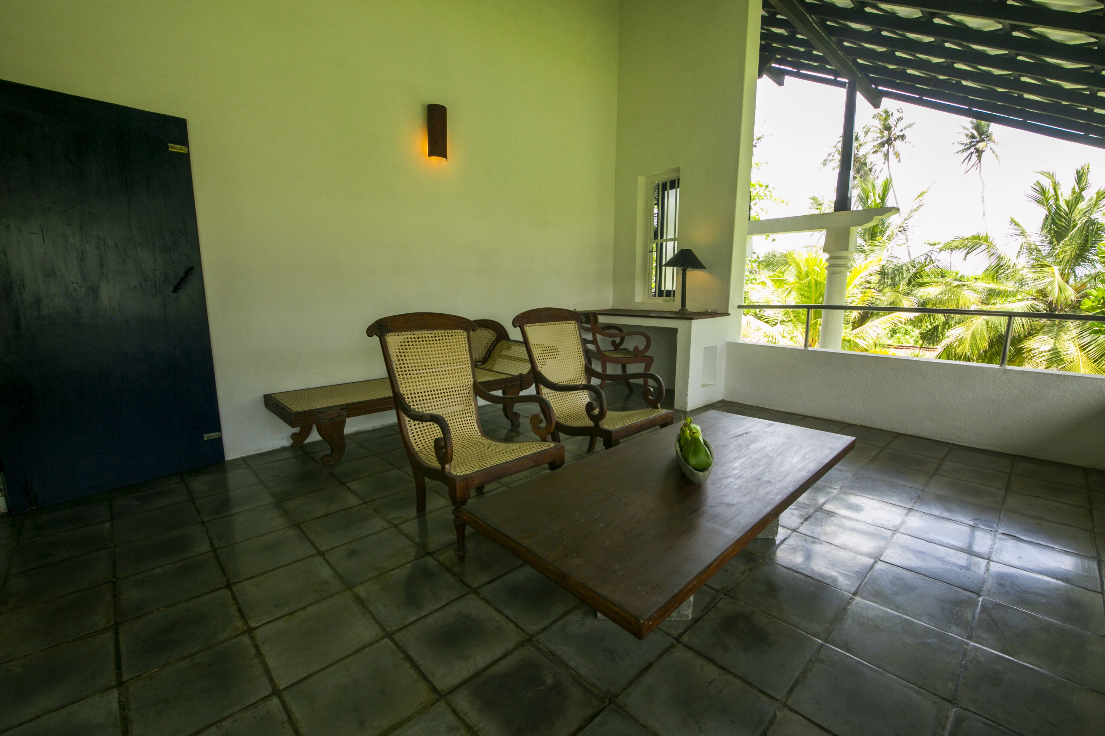 Una habitación con una mesa de madera, dos sillas de mimbre, un banco, una lámpara de pared y un ventanal con vista a palmeras.