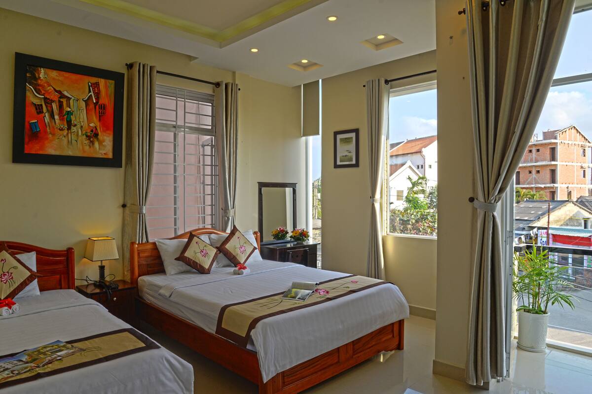 A hotel room with two beds, a large window, a painting on the wall, and a view of buildings outside.