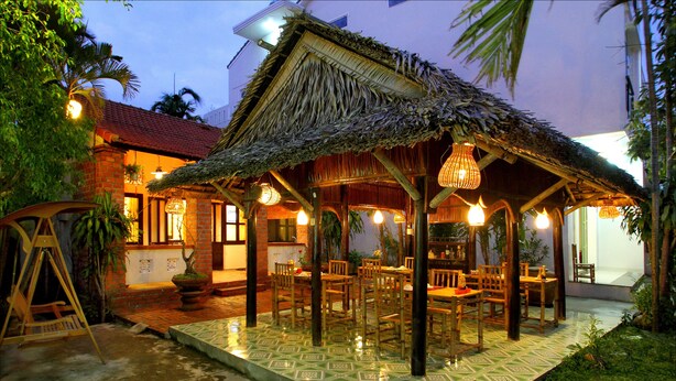 A thatched-roof outdoor dining area with wooden tables and chairs, surrounded by potted plants and a tiled floor. - Bamboo Garden Homestay (Da Nang)