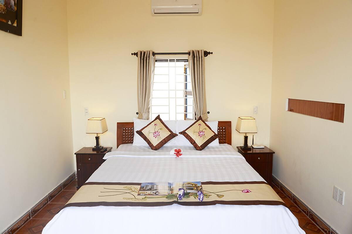 A hotel room with a large bed, two bedside tables with lamps, a window with curtains, and a wall-mounted air conditioner.