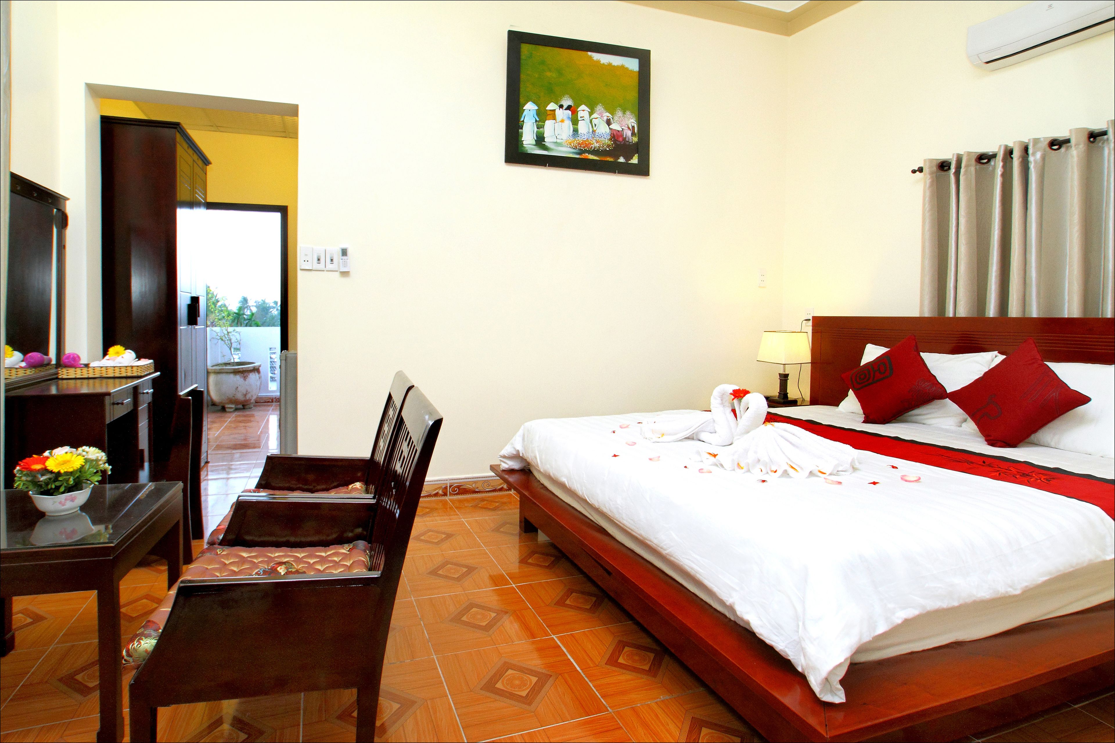 A hotel room with a bed, a desk, a chair, a small table with a flower vase, and a painting on the wall.