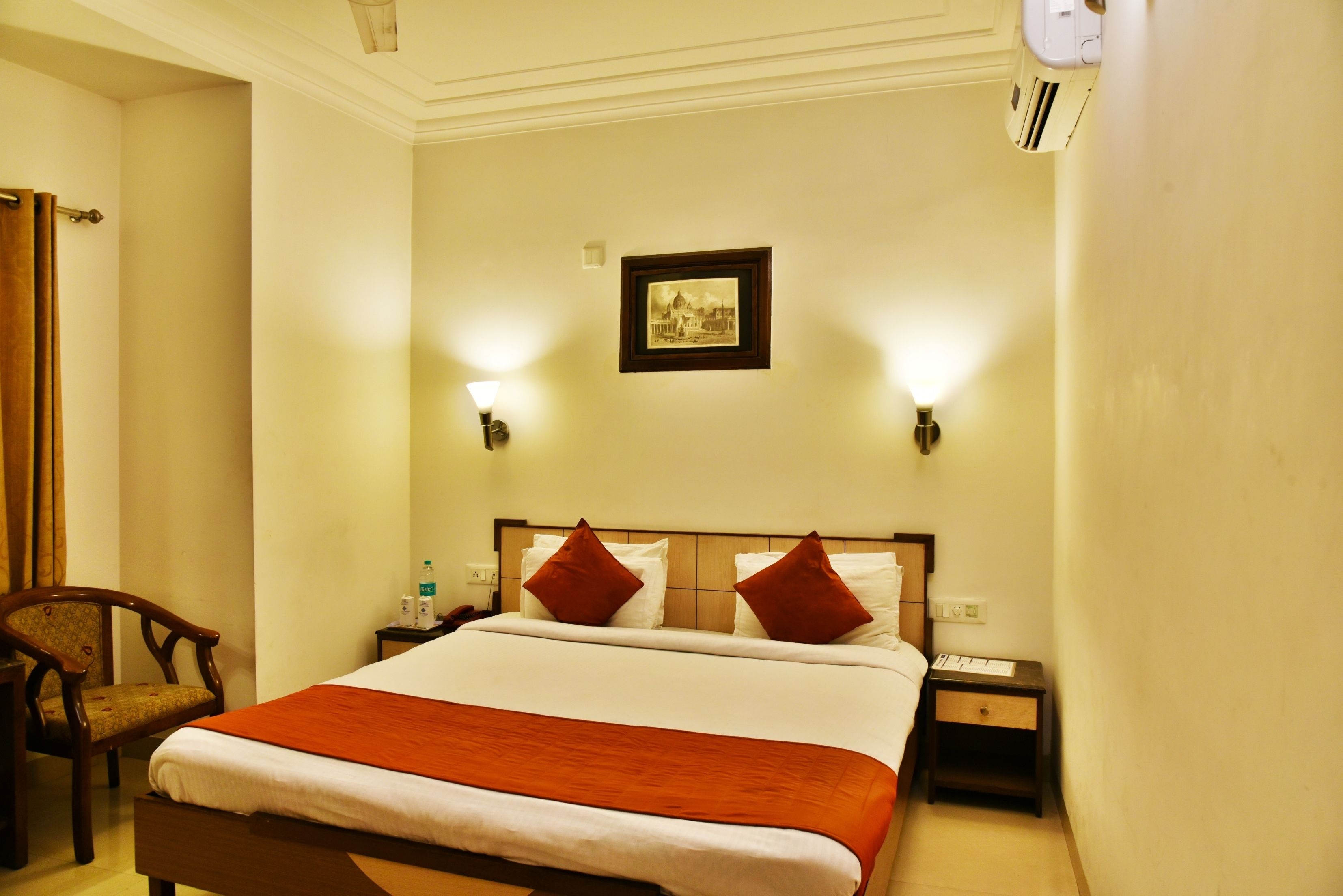 A hotel room with a bed, a chair, a nightstand, and a wall-mounted air conditioner.
