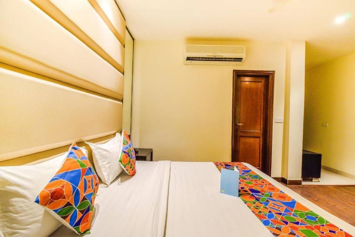 A hotel room with a bed, a wooden door, an air conditioning unit, and a small table with a lamp.