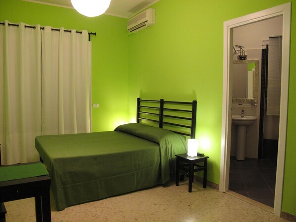 A room with a bed, a nightstand with a lamp, a bathroom visible through an open door, and an air conditioning unit on the wall. - Arches Inn B&B (Rome)