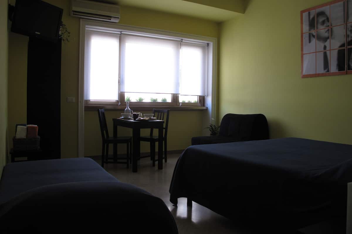 Una habitación con dos camas, una mesa con sillas, una ventana con persianas, un aire acondicionado y un cuadro colgado en la pared.