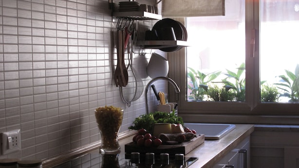 Cocina con salpicadero de azulejos blancos, una ventana con plantas y una encimera con un bol de frutas y verduras.