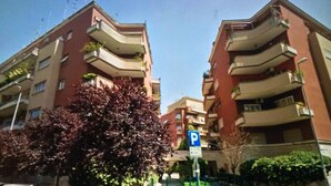 Un edificio residencial de varios pisos con balcones, un cartel de estacionamiento y un árbol con hojas moradas.
