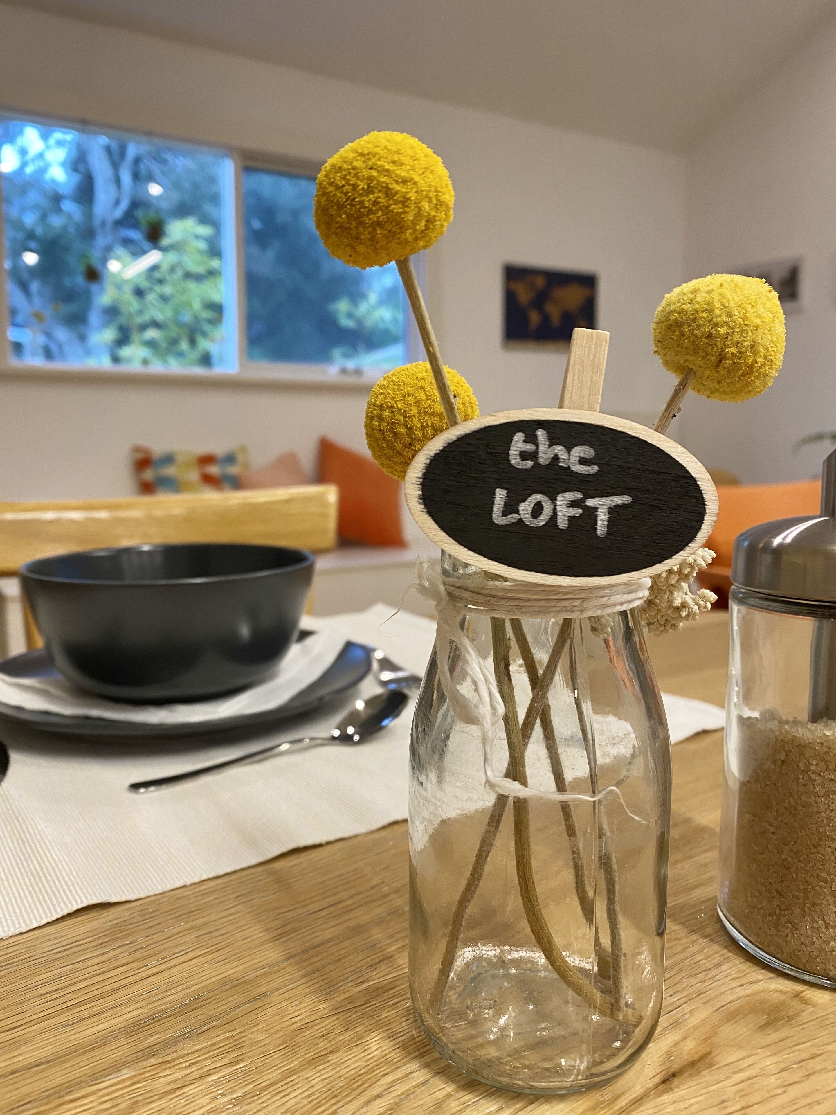 Un frasco de vidrio con una etiqueta que dice “the loft” y dos pompones amarillos en palitos, sobre una mesa de madera con un tazón y una cuchara al fondo.