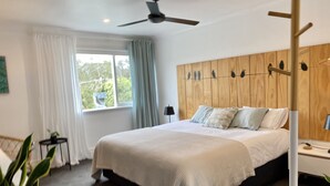 A modern bedroom with a large bed, a ceiling fan, a wooden headboard with bird decorations, a bedside table with a lamp, and a view of greenery outside. - Sunny Side Up B&B (Rye)