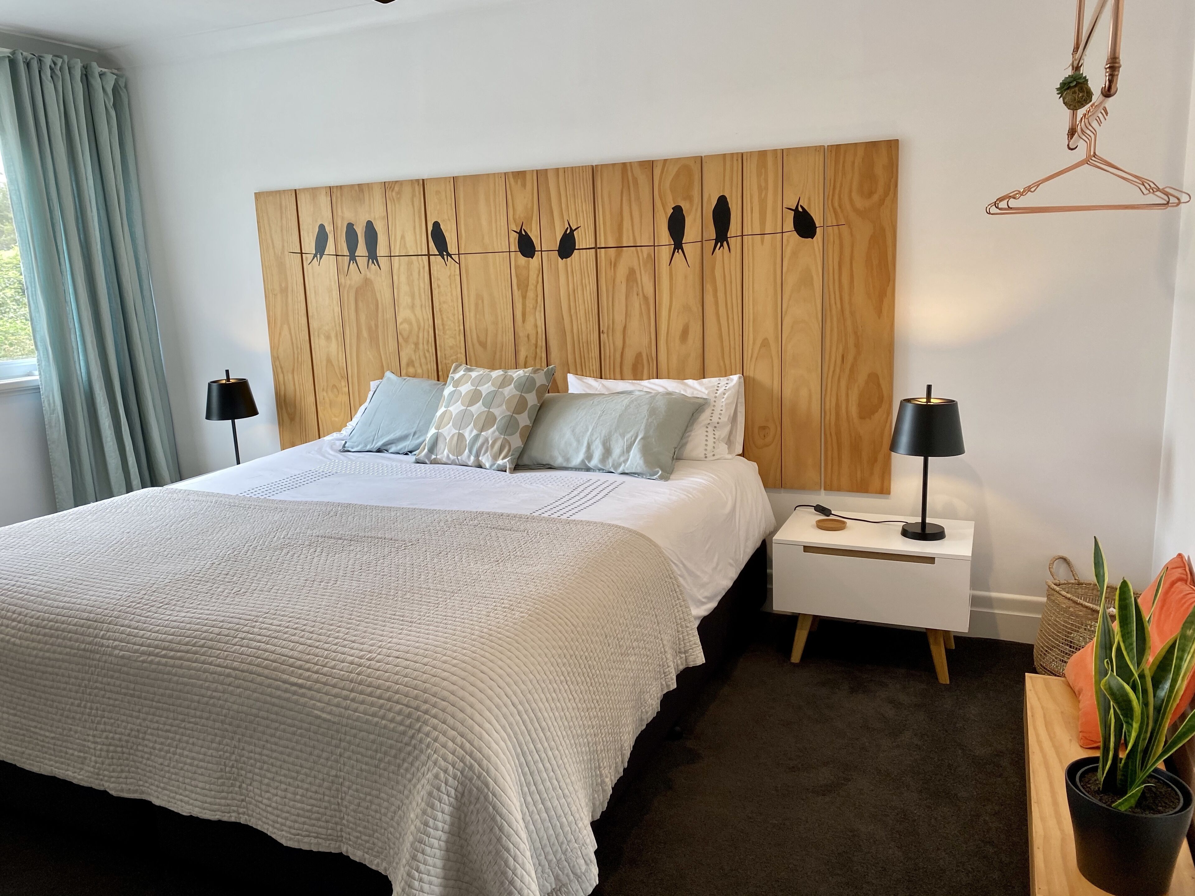 Ein ordentlich eingerichtetes Schlafzimmer mit einem Holzkopfende, einem großen Bett mit weißer Steppdecke, zwei Nachttischen mit Lampen und einer hängenden Pflanze.