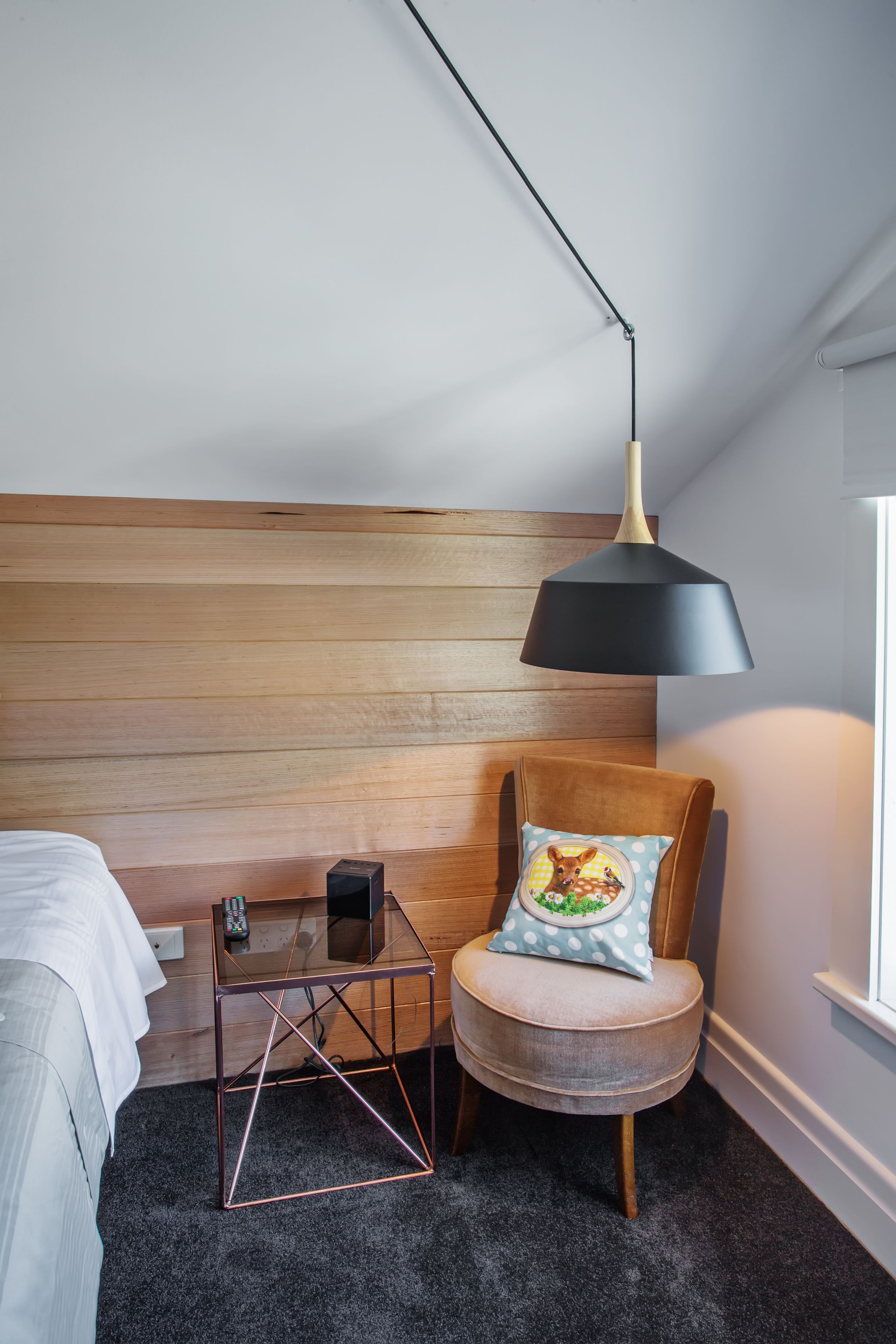 Een slaapkamer met een houten hoofdbord, een ronde stoel met kussen, een tafeltje met lamp en een raam.