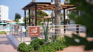 A café-bar named 'ASTRÁEAS' with a pergola structure, palm trees, and a pool area in the background. - Astreas Beach Hotel (Paralimni)