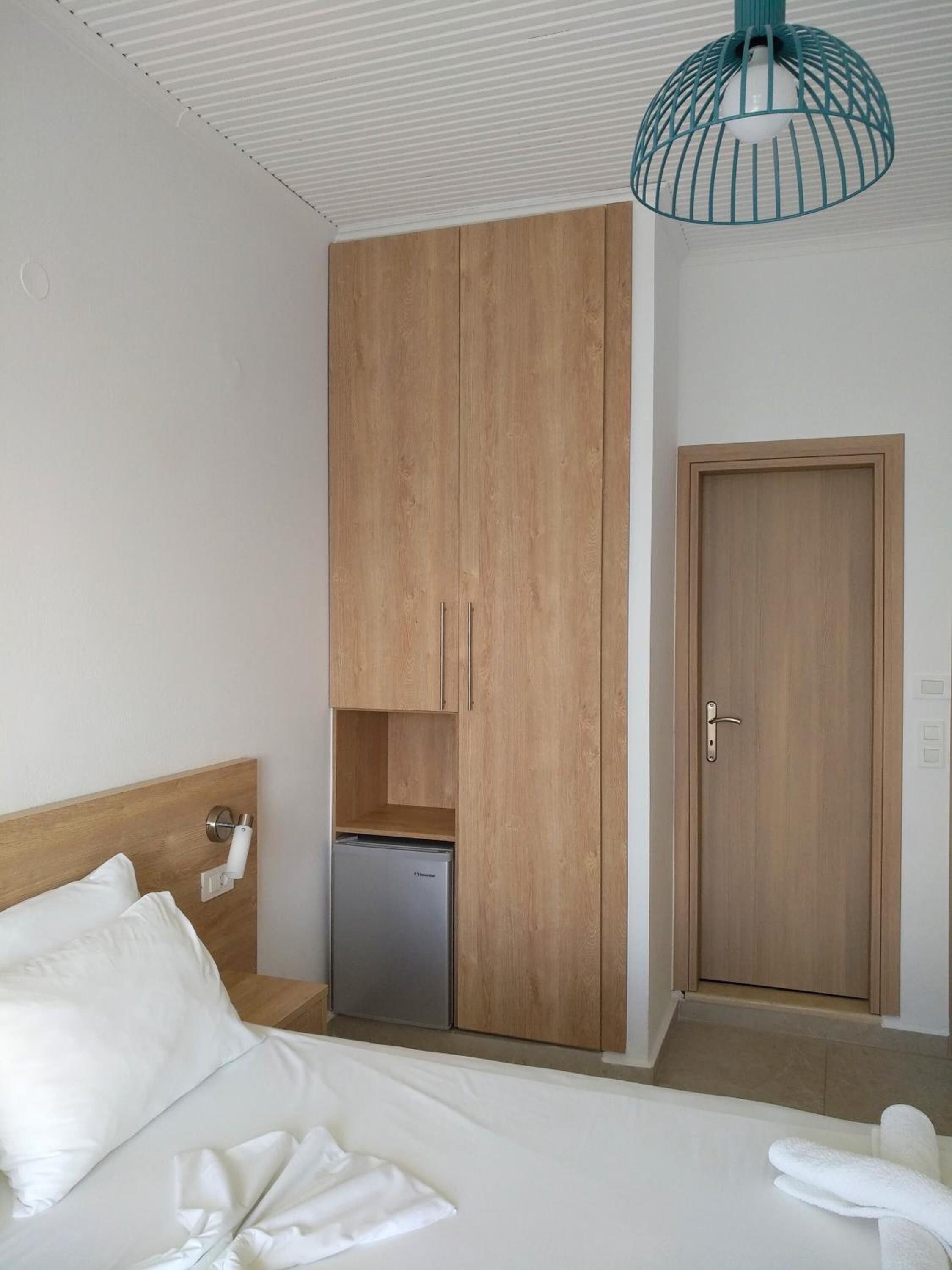 A hotel room with a bed, a wooden wardrobe, a small refrigerator, a door, and a hanging light fixture.
