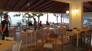 Zona de menjador interior amb taules i caires de fusta, un ventilador de sostre i vista al mar.