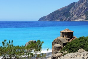 Un paisaje costero con mar azul cristalino, terreno rocoso y una pequeña construcción de piedra.