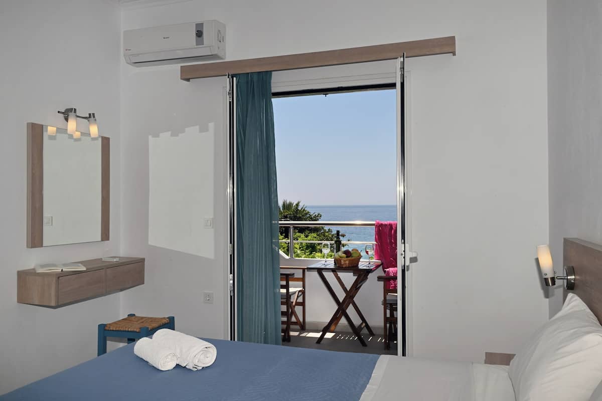 A hotel room with a bed, a small table with a basket of fruit, a chair, and a balcony with a view of the sea.