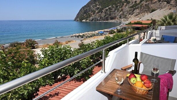 Un balcó amb taula plena de fruita, ampolla i copa, amb vista a una platja d'umbrel·les i un paisatge de muntanyes.