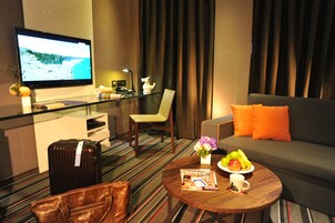 A hotel room with a television, a glass table with a magazine, a sofa with orange cushions, and a tray with fruit.