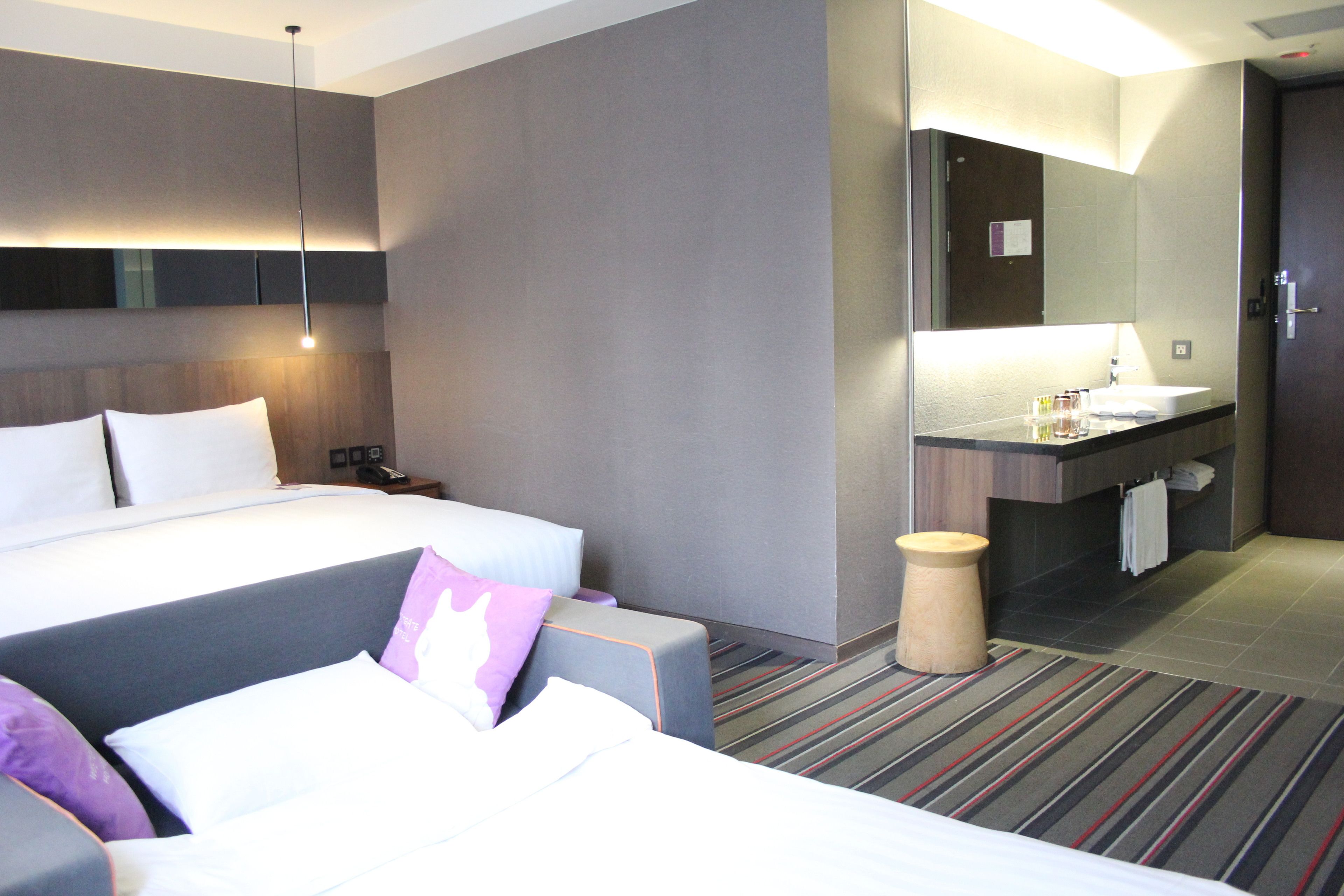 Habitación de hotel con una cama grande, una mesita de noche, un tocador, un baño con lavamanos y espejo, y una alfombra a rayas.