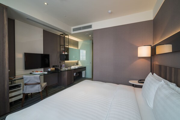 Una habitación de hotel moderna con una cama grande, un escritorio con televisor de pantalla plana, un baño con lavamanos y espejo, y una mesita con lámpara.