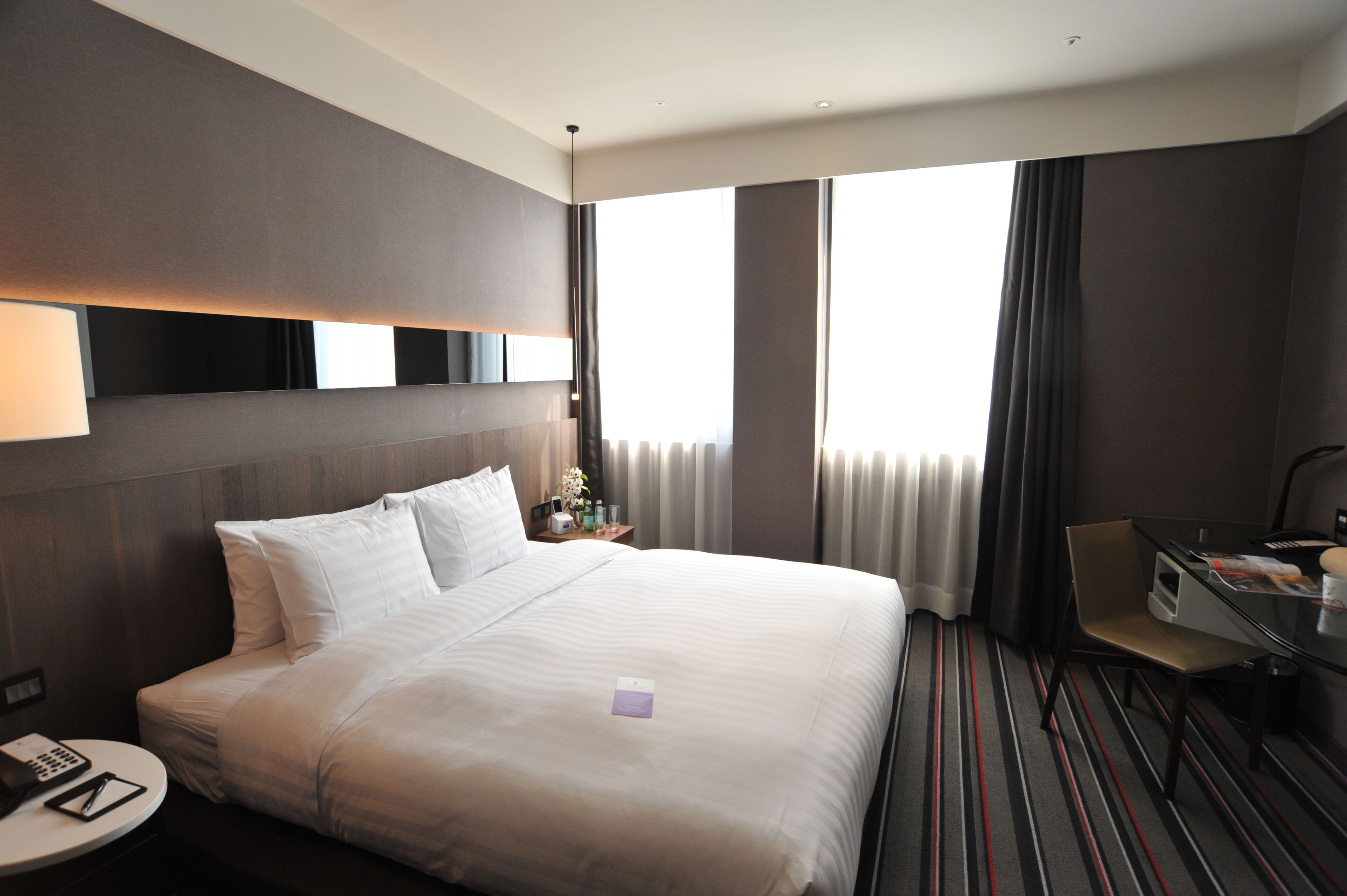 A modern hotel room with a large bed, a desk, and a view of the city through large windows.