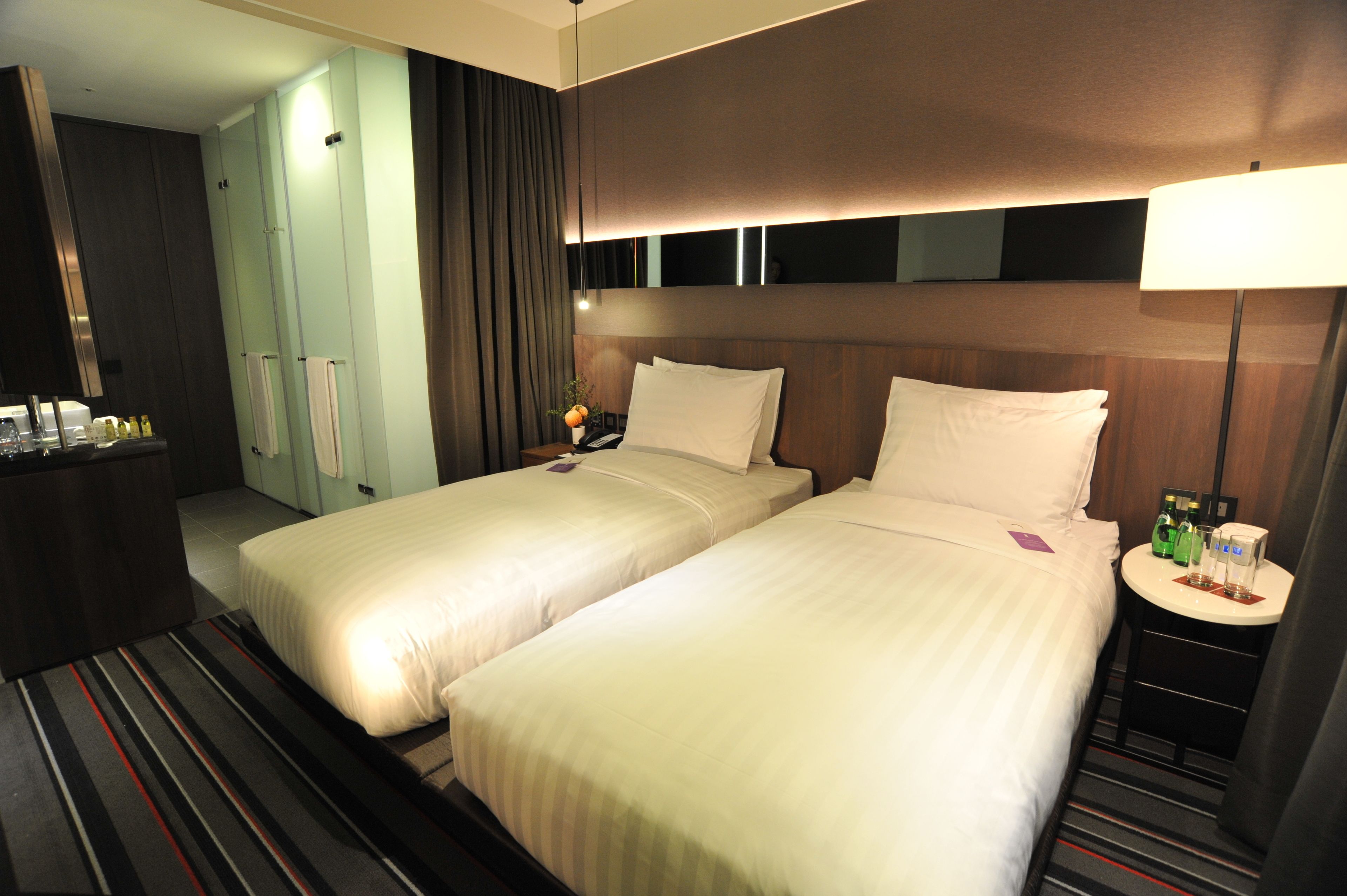 A hotel room with two beds, a small table with a lamp, a minibar, and a bathroom with a mirror and curtains.