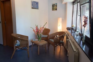 Quarto com móveis de vime, um vaso de flores e vista pela janela com cortinas leves.