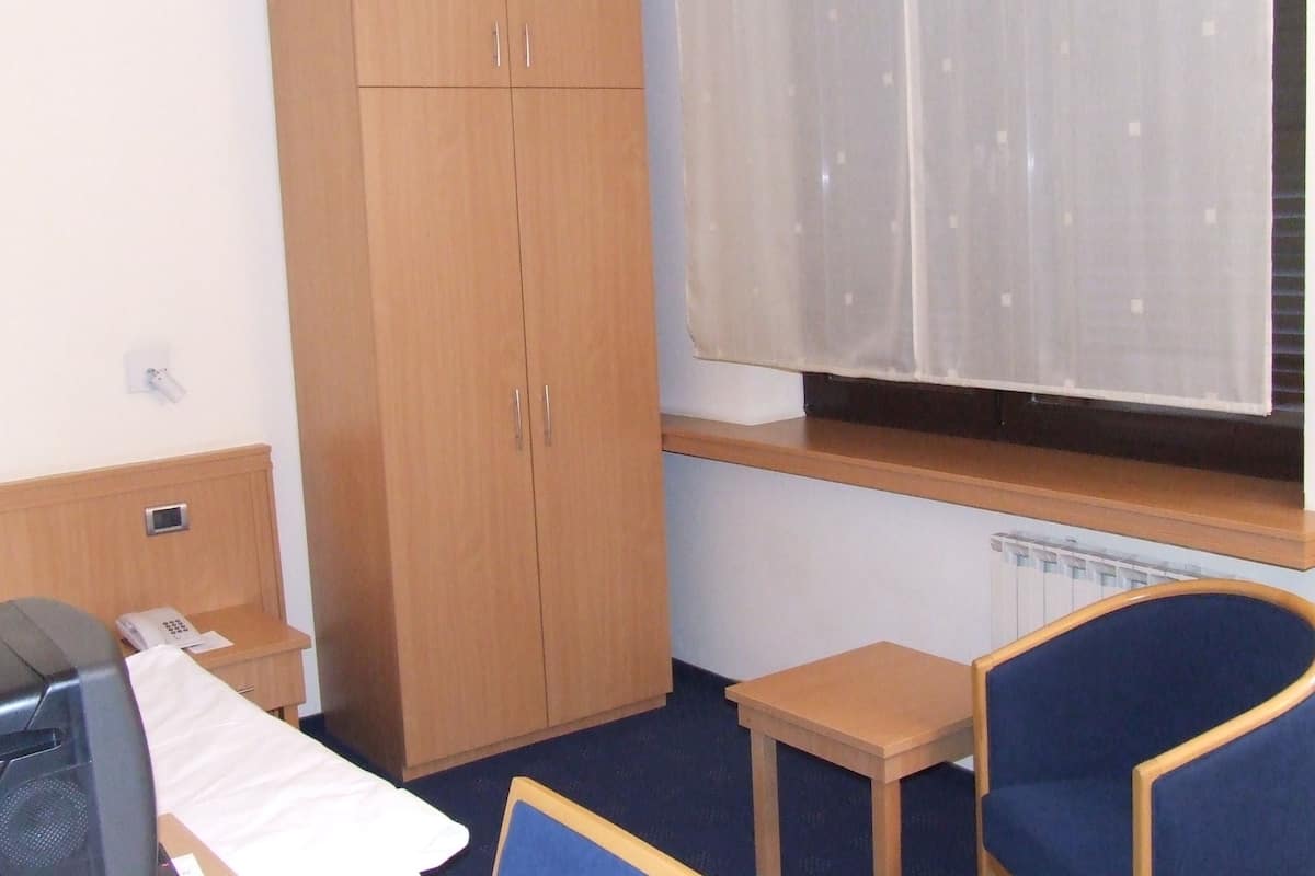 A small room with a wooden wardrobe, a small table, a blue chair, and a window with a sheer curtain.