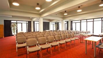 Una sala conferenze con file di sedie e tavoli, ampie finestre e luci a soffitto.