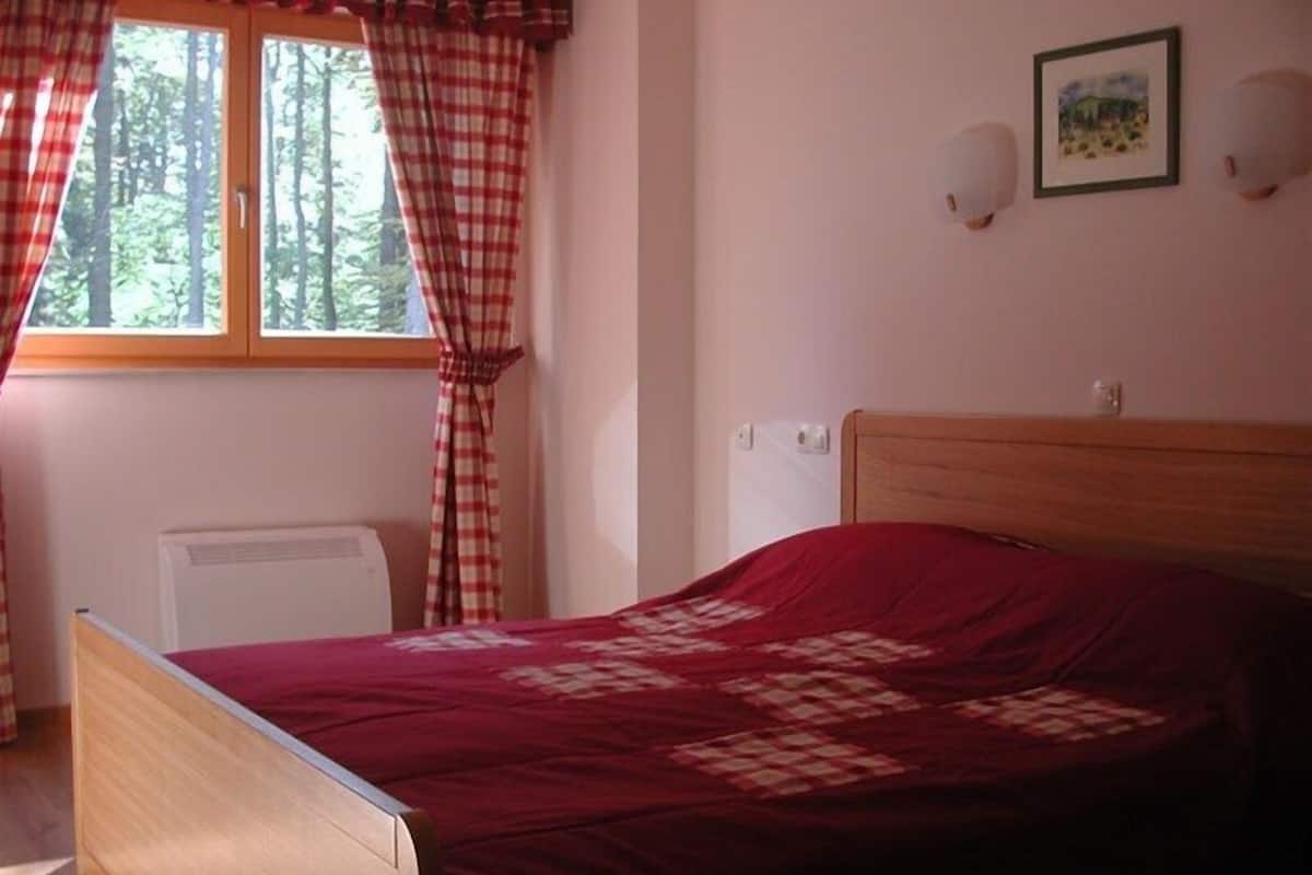 A bedroom with a wooden bed, a window with checkered curtains, a radiator, and a framed picture on the wall.