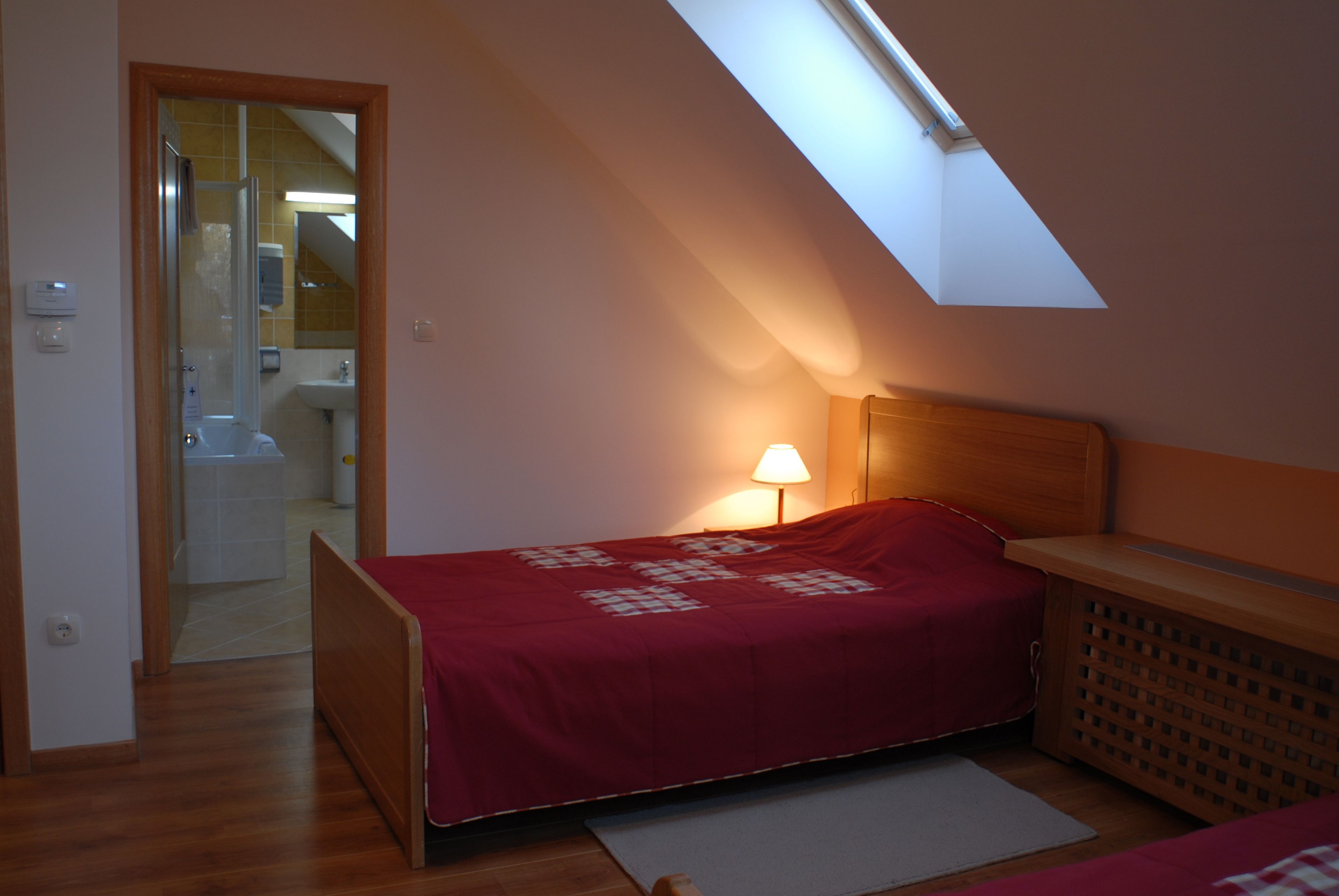Une chambre avec un puits de lumière, un lit recouvert d’une couverture rouge, une table de chevet et une tête de lit en bois.