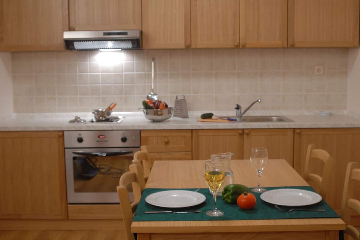 A kitchen with wooden cabinets, a stove, and a dining table set with plates, a glass of wine, and vegetables.