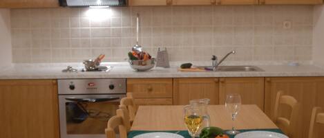 A kitchen with wooden cabinets, a stove, and a dining table set with plates, a glass of wine, and vegetables.