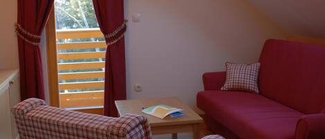 A cozy room with a red sofa, a checkered armchair, and a small table with a book. A window with red curtains and a wooden blind is visible.