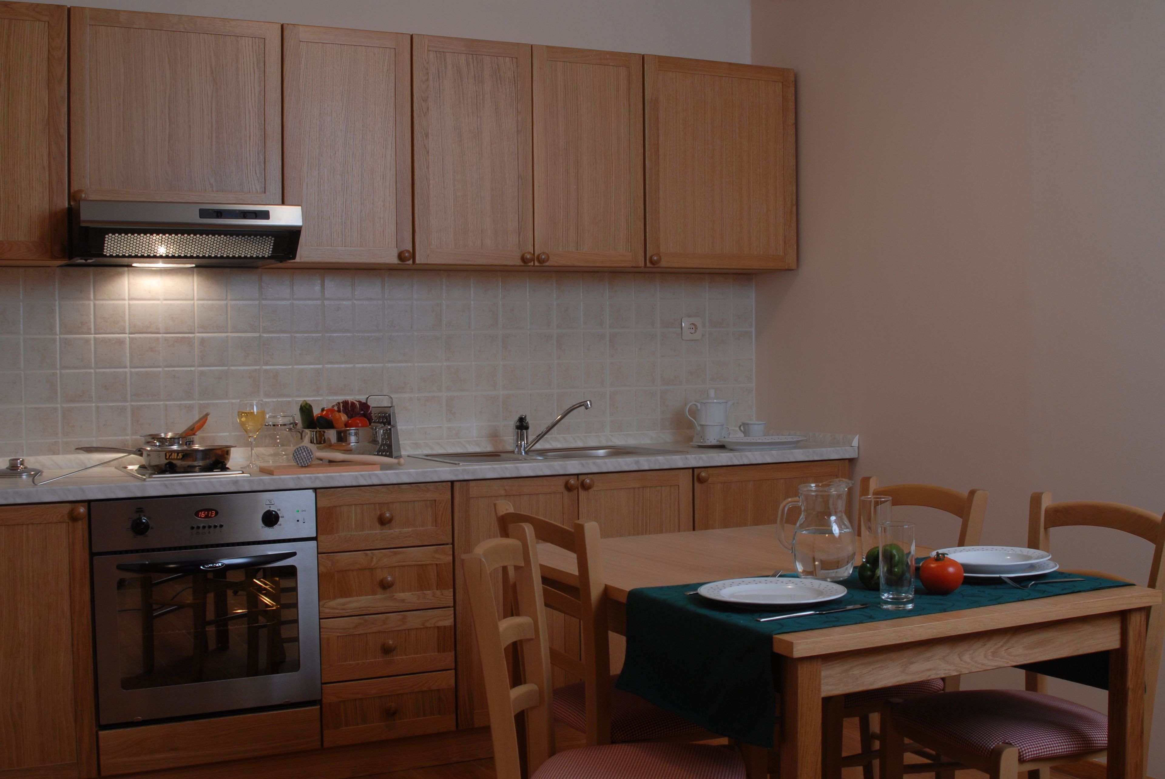 Een keuken met houten kastjes, een roestvrijstalen oven, een spoelbak en een eettafel gedekt met borden en een karaf.