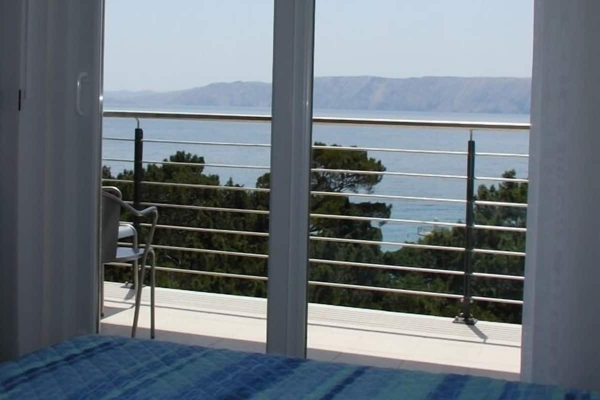 A bedroom with a bed, a chair, and a balcony with a view of the sea and mountains.