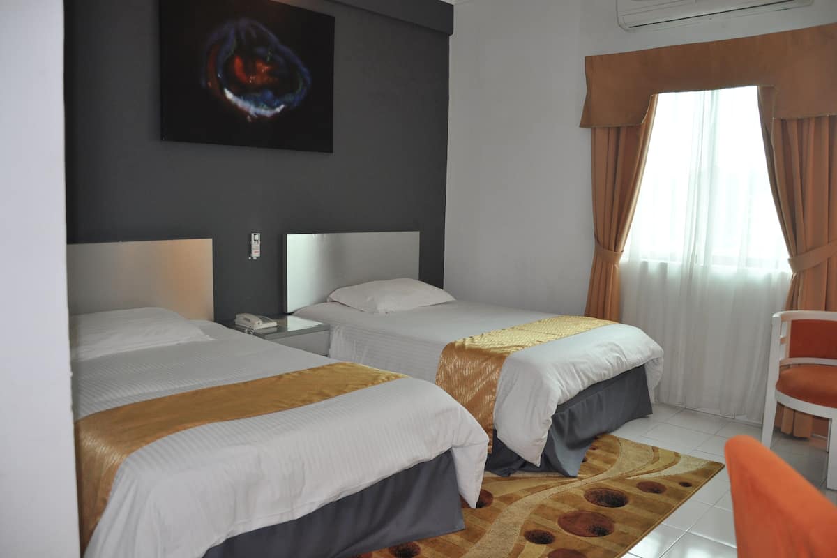 Een hotelkamer met twee bedden, een bureau met telefoon, een stoel, een schilderij aan de muur en een raam met gordijnen.